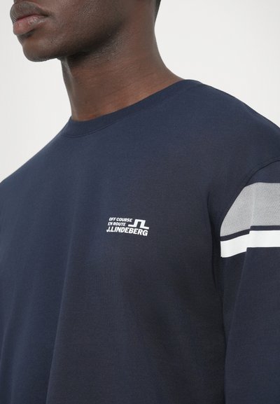 Μπλε σκούρο βαμβακερό μπλουζάκι με κοντά μανίκια, διακοσμημένο με λευκό λογότυπο στο στήθος και γκρι και λευκές ρίγες στα μανίκια.