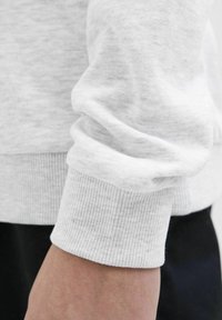 Tessuto di un maglione grigio chiaro con una texture morbida, caratterizzato da polsini a coste e una vestibilità rilassata, mostrato dalla parte centrale del braccio fino al polso.