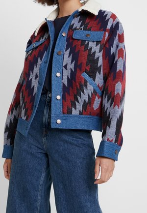 Osoba ubrana w kurtkę z kolorowym wzorem zygzaka, z akcentami dżinsu, kołnierzem z owczej skóry oraz niebieskie, wysokowodne jeansy.