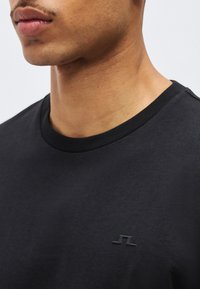 Κοντινό πλάνο λαιμού και κάτω μέρους προσώπου άνδρα που φοράει μαύρο μπλουζάκι με στρογγυλή λαιμόκοψη και μικρό ανάγλυφο λογότυπο στο στήθος.