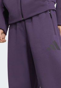 Violeta sporta treniņtērps, kas sastāv no jaku ar rāvējslēdzēju un biksēm. Audums ir gluds, ar matētu apdari un melnu logotipa detaļu uz biksēm.