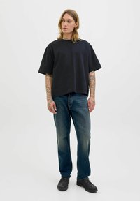 Zwarte oversized katoenen t-shirt, rechte blauwe spijkerbroek en zwarte schoenen. Model heeft lang haar en zichtbare tatoeages op de armen.