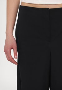 Black pants made of soft fabric with a smooth texture, featuring a flat front waistband and a streamlined silhouette. Hand resting at the waist.