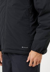 Man wearing black Snow Peak jacket and pants with visible zipper and elastic wrist cuffs, hand relaxed by side.