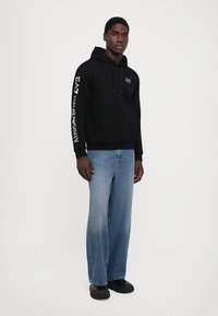 Felpa nera in cotone con tasca frontale, cappuccio con coulisse e logo EA7 sulla manica, abbinata a jeans larghi azzurri e scarpe nere.