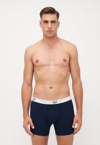 Boxeri scurți de culoare bleu marin, cu un elastic alb pe talie, având logo-ul 'GAP'. Material neted, design ajustat și lungime până la mijlocul coapsei.
