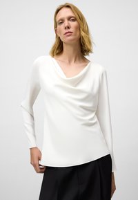 Top bianco a maniche lunghe con scollo a drappeggio, texture liscia e orlo arrotondato, abbinato a pantaloni scuri.