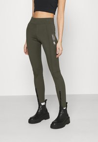 Leggings verde oliva en una tela elástica con una cintura alta, diseño elegante y acentos con cremallera en los tobillos. Detalle del logo en el lateral.