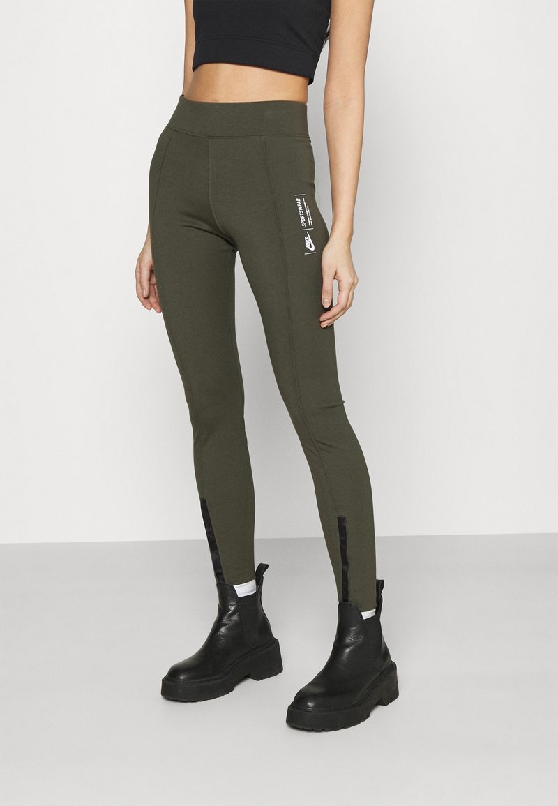 Leggings verde oliva en una tela elástica con una cintura alta, diseño elegante y acentos con cremallera en los tobillos. Detalle del logo en el lateral.