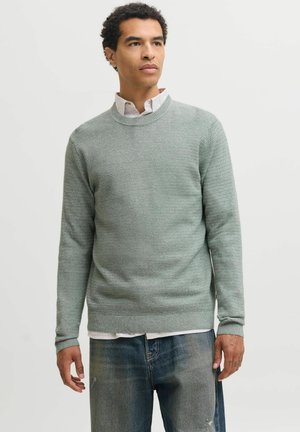 Jeune homme portant un pull gris clair texturé par-dessus une chemise blanche à col et un jean bleu délavé, debout devant un fond clair uni.