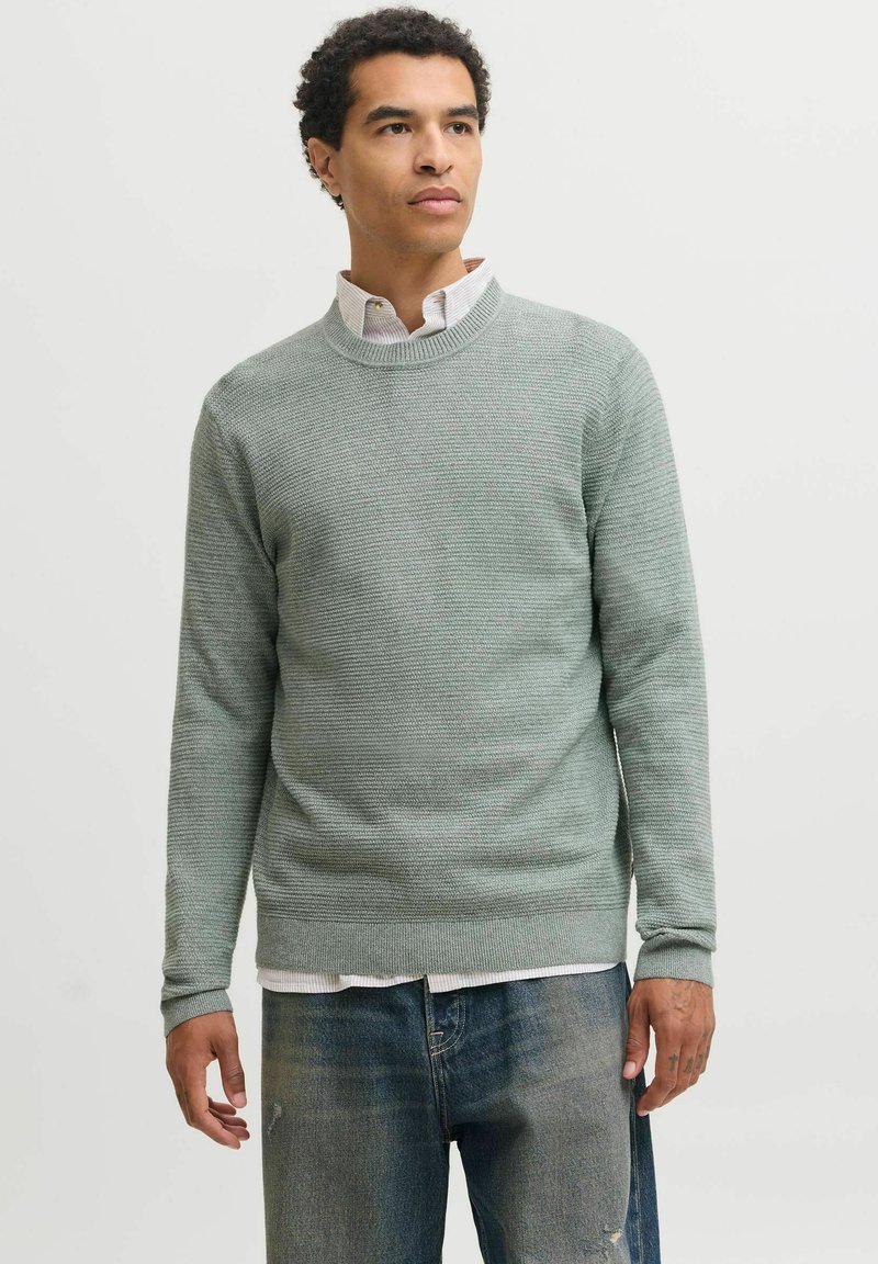 Jeune homme portant un pull gris clair texturé par-dessus une chemise blanche à col et un jean bleu délavé, debout devant un fond clair uni.