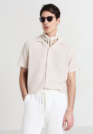 Beige kurzärmliges Oberhemd mit Struktur und Camp-Kragen, kombiniert mit cremafarbigen Jogginghosen mit Kordelzug, und accessoriert mit einem leichten Schal und dunklen Sonnenbrillen.