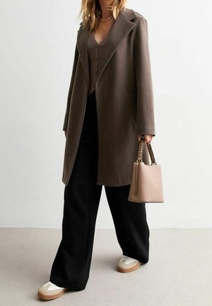 Kobieta ubrana w długi brązowy płaszcz, czarne spodnie o szerokich nogawkach, białe trampki i trzymająca beżową torebkę z plecionymi rączkami.