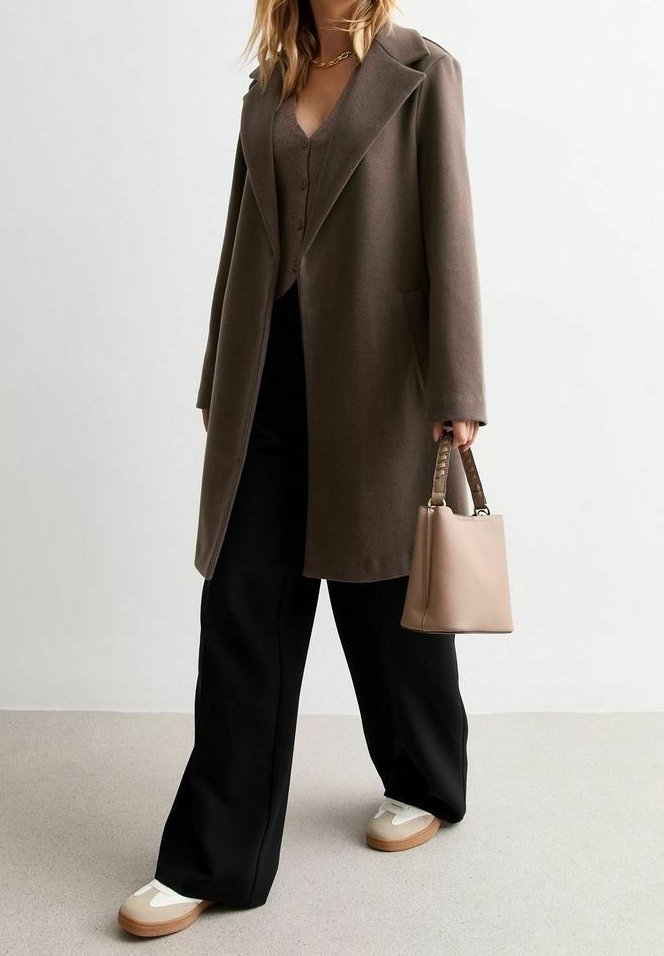 Femme portant un long manteau marron, un pantalon large noir, des baskets blanches, et tenant un sac à main beige avec des anses tressées.