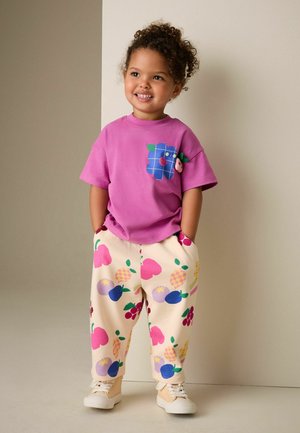 Kleinkind mit lockigem Haar lächelt, trägt ein violettes Shirt mit einem blauen Blumenaufnäher und cremefarbene Hose, die mit bunten Obstmustern verziert ist.