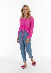 Rosa offenstrickter Pullover mit Zugbanddetails, kombiniert mit hellblauen, umgeschlagenen Jeans und rosa Slides mit dekorativen Akzenten.