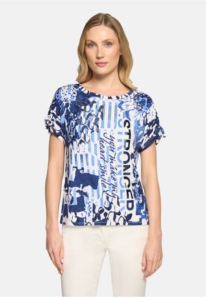 Femme blonde portant une blouse à manches courtes bleue et blanche à motifs avec du texte, associée à un pantalon clair, debout devant un fond blanc.