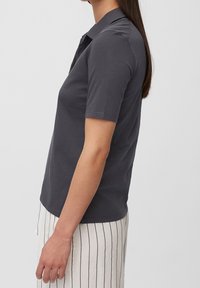 Kvinna iklädd en mörkgrå kortärmad polo och vita byxor med vertikala svarta ränder, stående på sida mot en vit bakgrund.