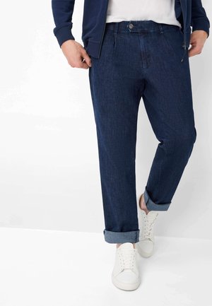 Mand iført mørkeblå opbukte jeans, hvide sneakers, hvid t-shirt og navyblå lynlåstrøje, stående med det ene ben fremad.