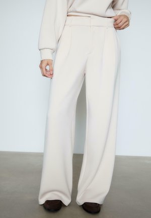 Personne portant un pantalon crème taille haute à jambes larges associé à un pull crème assorti et des chaussures marron foncé, debout sur un sol en béton.