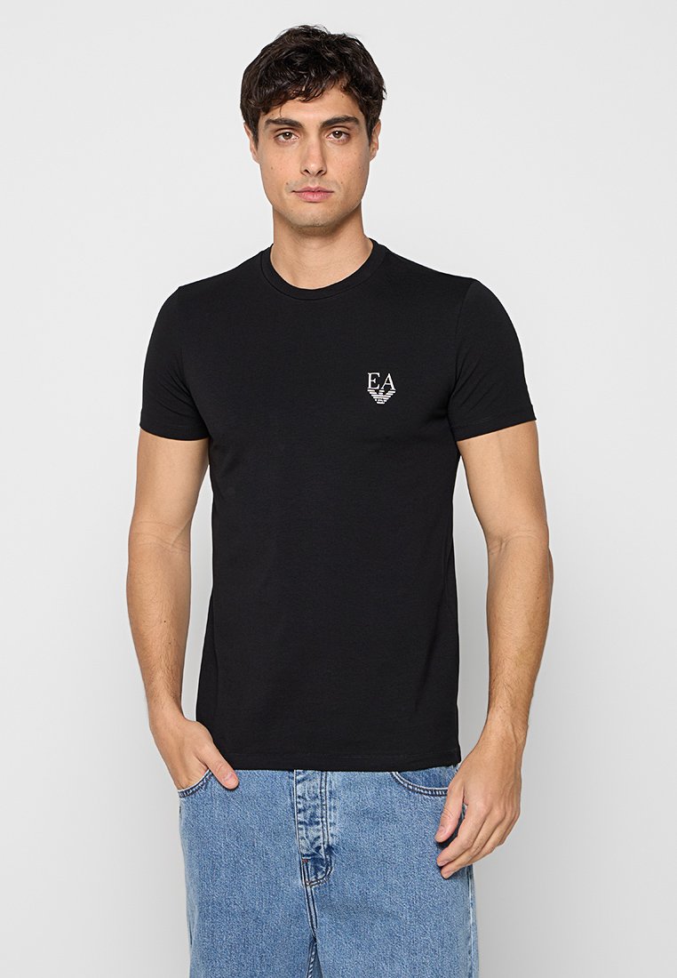 Hombre vistiendo una camiseta negra ajustada con el logo "EA" en el pecho y vaqueros azul claro, de pie frente a un fondo neutro.