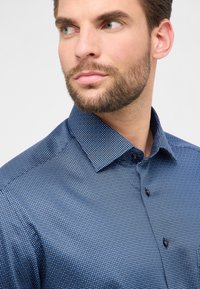 Camicia blu con motivo a pois, dotata di colletto e chiusura con bottoni. Il tessuto presenta una leggera texture.