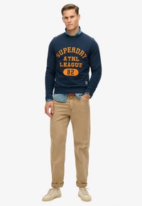 Sudadera azul marina con estampado naranja "Superdry ATHL League 82", superpuesta sobre una camisa de mezclilla clara, combinada con pantalones beige y zapatillas crema.