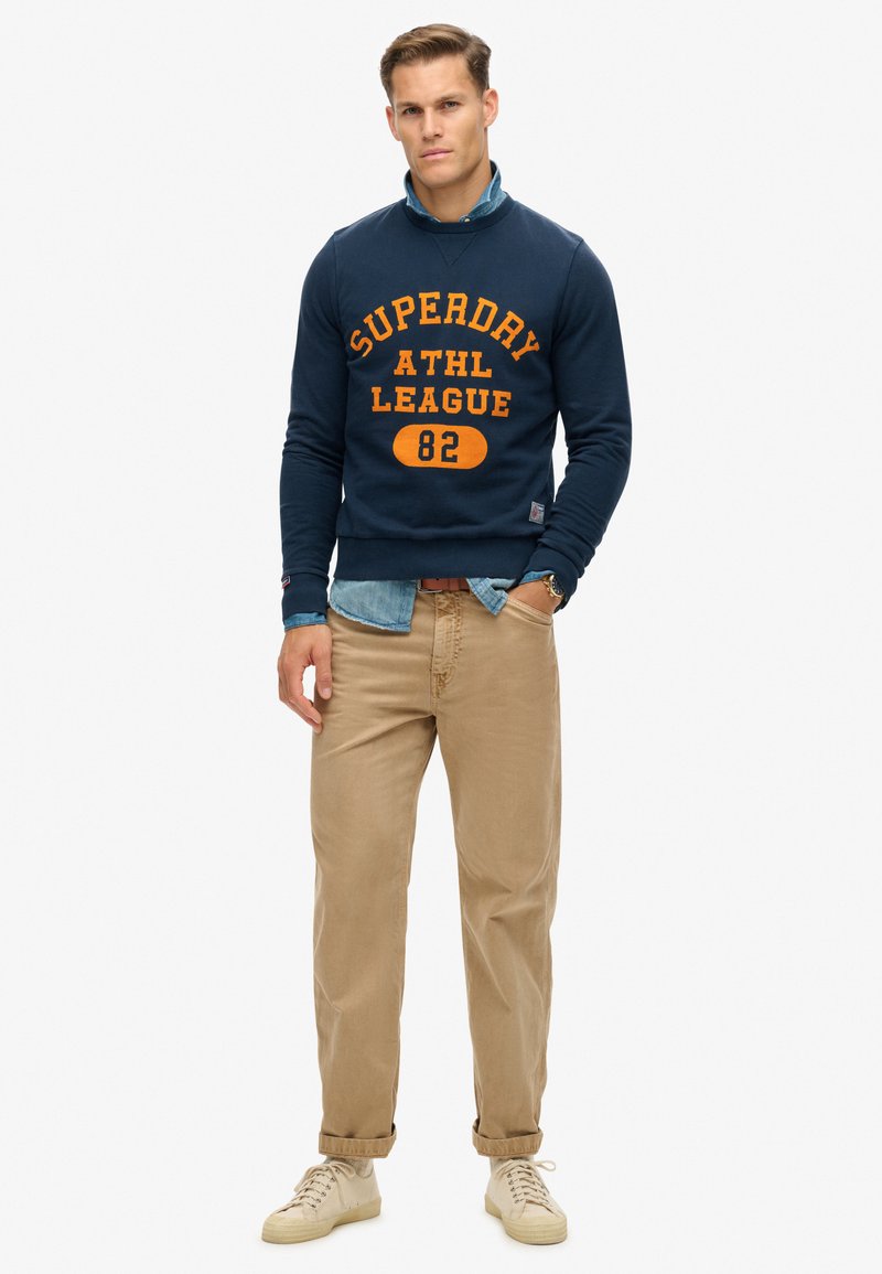 Marineblauer Sweatshirt mit orangefarbener "Superdry ATHL League 82" Aufschrift, über einem hellen Denimhemd getragen, kombiniert mit beigen Hosen und cremefarbenen Sneakern.