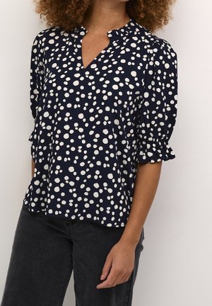 Woman wearing a navy blouse with white irregular polka dots and ruched short sleeves, paired with dark gray jeans.