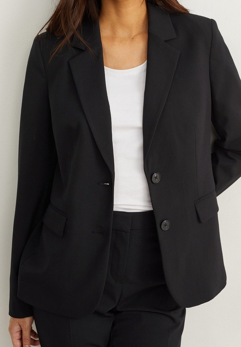 Femme portant un blazer noir cintré sur un haut blanc et un pantalon noir assorti, se tenant devant un fond uni.