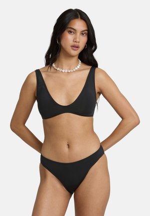 Kobieta w czarnym komplecie bikini i naszyjniku z pereł, stojąca z rękami za plecami na jednolitym białym tle.