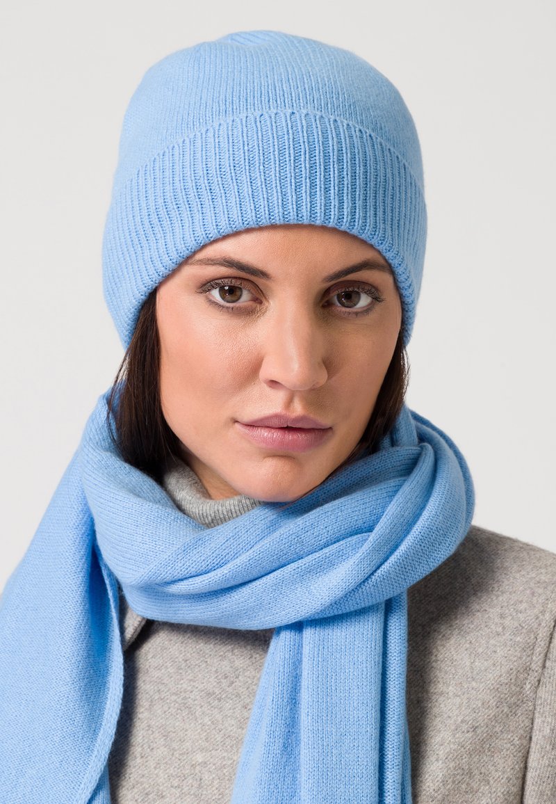 Bonnet en tricot bleu clair avec une texture côtelée, associé à un foulard doux assorti. L'arrière-plan est gris neutre, mettant en valeur les accessoires.