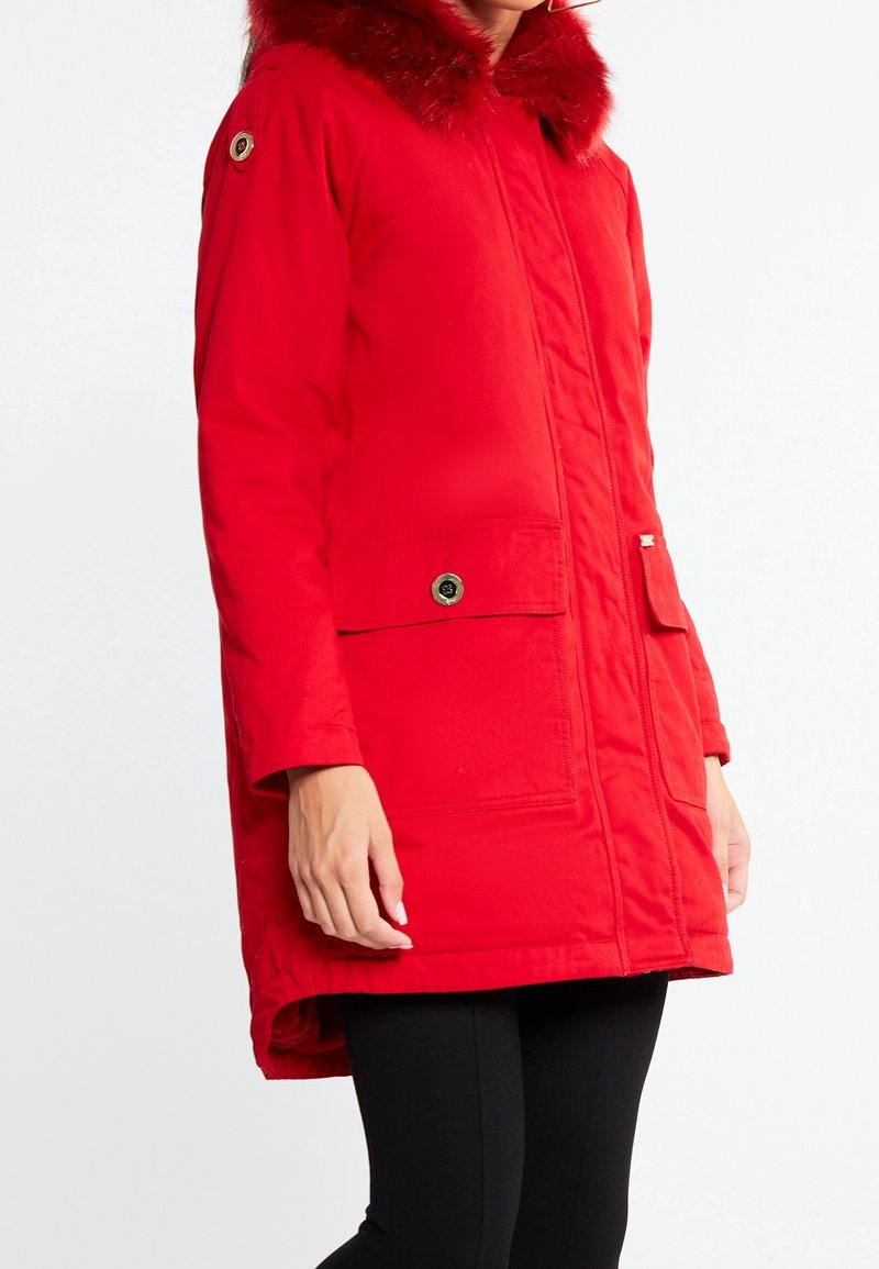 Parka rouge isolante avec capuche en fausse fourrure, poches avant et fermeture éclair. Présente une coupe droite et une texture douce.