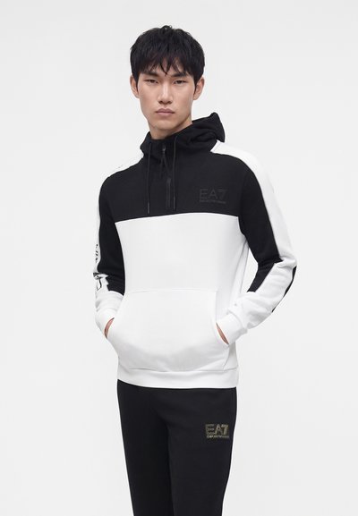Млад мъж, облечен в черно-бял суитшърт EA7 Emporio Armani и черен панталон с лого, с ръце в джобовете, гледащ напред на едноцветен фон.