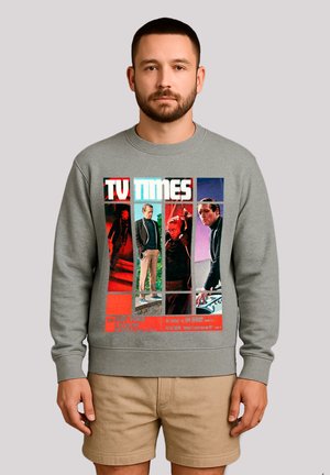 Mann in grauem Sweatshirt mit buntem "TV TIMES" Magazincover, das vier Männer in verschiedenen Posen zeigt, kombiniert mit beigen Shorts.