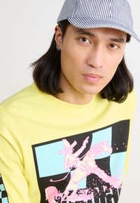 T-shirt en coton jaune avec un design graphique noir et bleu représentant un cow-boy rose sur un cheval. Une casquette à rayures bleues et blanches complète le look.