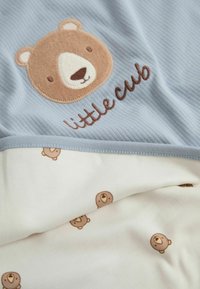 Világoskék anyag, amelyen egy medve arcának hímzése és a "little cub" felirat látható. Az alatta lévő fehér anyagon ismétlődő, kis medve minta található. Puha tapintású.