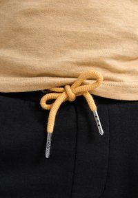 T-shirt beige en tricot rentré dans un pantalon noir, avec un cordon tressé jaune et des embouts en argent noués à la taille.