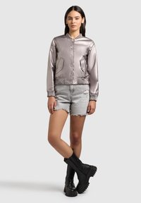 Femme portant une veste bomber métallique lavande, un short en jean gris clair effilé, et des bottes de combat noires à lacets, debout, jambes croisées.