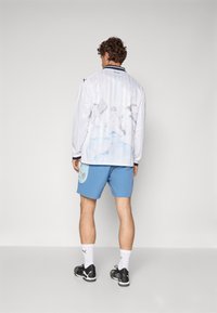 Camiseta blanca de manga larga con patrones en acuarela azul, combinada con pantalones cortos azules que presentan un parche. Medias largas y zapatillas negras completan el atuendo.