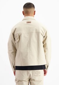 Beige jas van een gestructureerde stof, met knoopsluiting en een logo-label op de achterkant. Het ontwerp heeft een losse pasvorm en zakken.