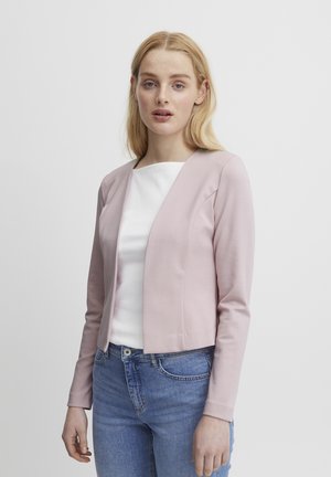 Veste courte rose clair en tissu doux avec un col rond et des manches longues, associée à un top blanc et un jean bleu.