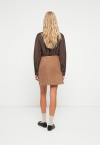 Bruine blouse met lange mouwen en een gathered taille, gecombineerd met een tan suède rok. Model draagt zwarte ballerina's en beige sokken. Neutrale achtergrond.