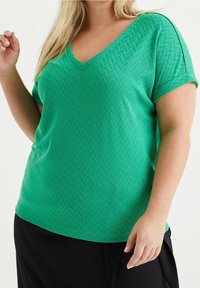 Grön kortärmad V-ringad topp med en texturerad herringbone-mönstring. Designad med en avslappnad passform och mjukt tyg för komfort.