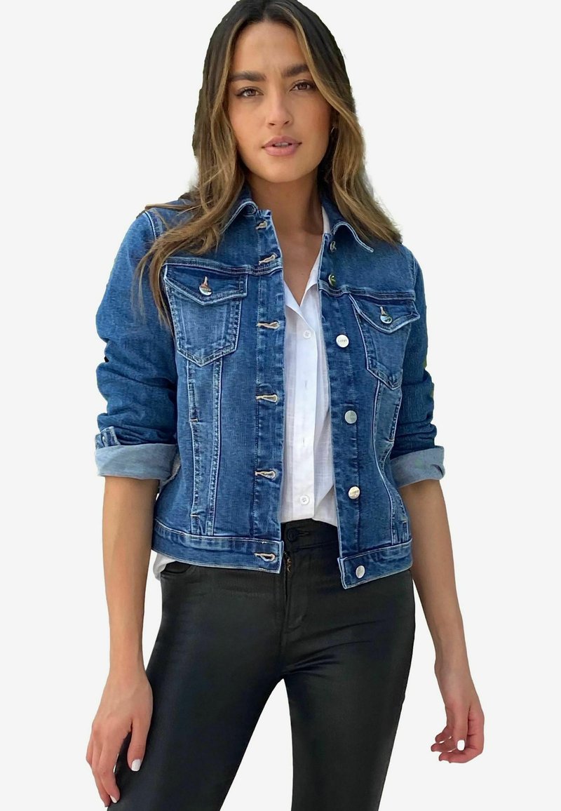 Donkere blauwe spijkerjack met knoopsluiting aan de voorkant, twee borstzakken en opgerolde mouwen, gecombineerd met een wit shirt en zwarte broek.