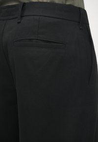 Pantalon noir sur mesure avec une texture lisse, comprenant deux poches arrière et une ceinture ajustée. Lignes épurées avec un design professionnel.