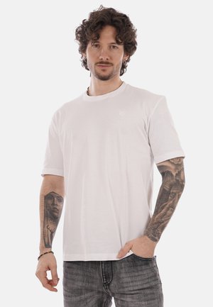 Uomo con capelli ricci che indossa una maglietta bianca semplice e jeans grigi, mostrando tatuaggi dettagliati su entrambi gli avambracci.