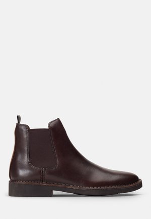 Bottine Chelsea en cuir marron avec côtés élastiques, bout arrondi et talon empilé. Présente des coutures le long de la semelle pour plus de durabilité.