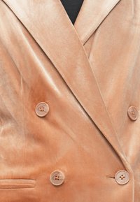 Gros plan sur un blazer en velours pêche à double boutonnage avec quatre grands boutons assortis et un col cranté sur un top noir.