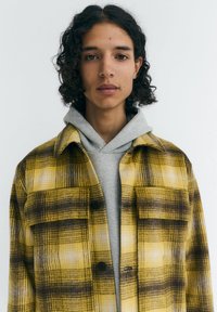 Gelb-braune karierte Hemd mit zwei vorderen Taschen und schwarzen Knöpfen, über einem grauen Kapuzenpullover mit Kordelzug getragen. Glatte Textur.